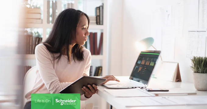 Eine Frau arbeitet an einem Schreibtisch an einem Laptop, auf dem Grafiken angezeigt werden, und hält ein Tablet in der Hand.