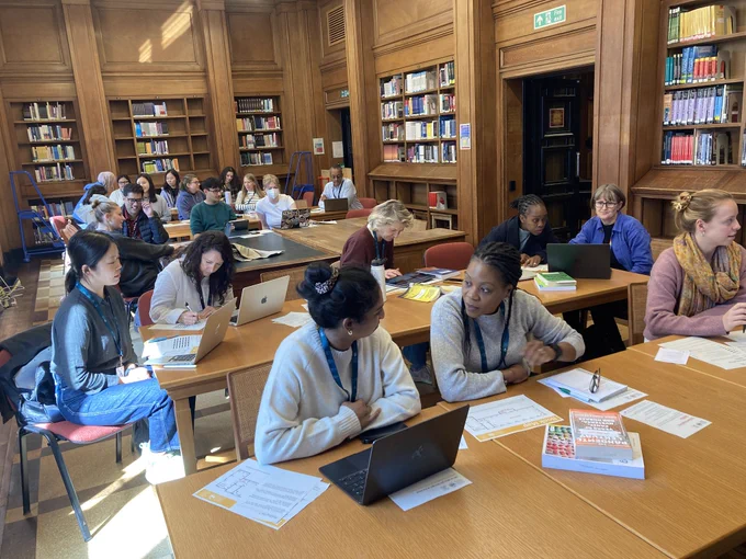 New students attending an induction session in the Barnard Room of the Library. Most are discussing something in pairs and many have laptops, books and other materials in front of them.