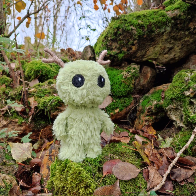 Photo en extérieur d'une peluche au crochet, elle a été faite avec du fil fourrure donc on dirait une poche cousue, c'est un petit personnage vert pastel de forme humanoïde dans un style assez chibi / mignon, avec juste des grand yeux noirs brodés et des petites cornes marrons. Les autres photos sont juste des angles différents 