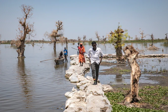 Three people walk along sandbags surrounded by water and submerged trees. A small boat is in the water alongside.