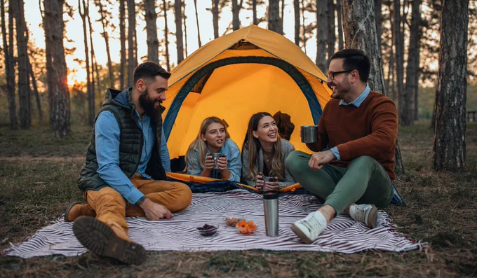Four adults enjoying snacks and drinks outside a yellow tent in a forest at sunset.