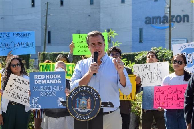 Senator Gianaris wears a light blue button down shirt and speaks at a podium with the seal of the New York State Senate. Behind him, advocates hold signs calling for cleaner air and accountability for polluters like Amazon, whose warehouse is visible in the background