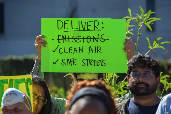 A green poster held aloft reads "Deliver: -Emissions (crossed out) - Clean Air - Safe Streets"