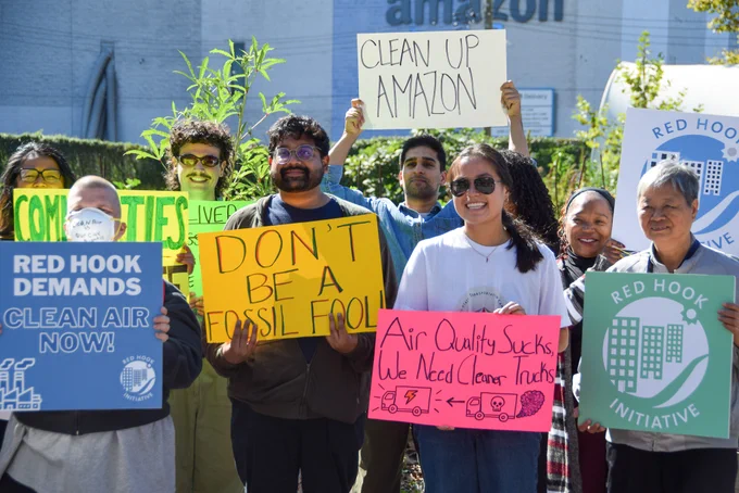 Advocates for clean air hold signs reading "Red Hook Demands Clean Air Now!", "Don't Be A Fossil Fool!", "Air Quality Sucks, We Need Cleaner Trucks", "Clean Up Amazon", and "Red Hook Initiative"