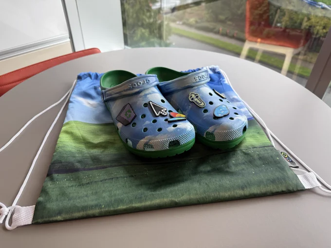Windows XP-inspired Crocs with custom Jibbitz charms sitting on top of a Bliss-themed drawstring backpack on a table.