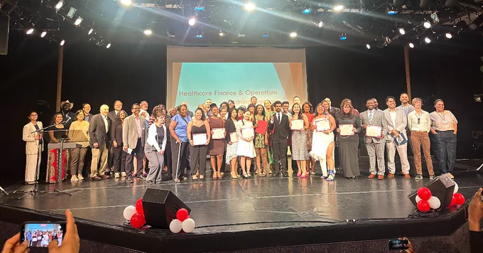S B S Commissioner Dynishal Gross and a large group of Healthcare Finance & Operations program graduates standing on stage holding their certificates at the graduation ceremony