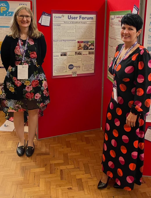 2 ladies standing in from of a poster board, a User Forum poster is visible