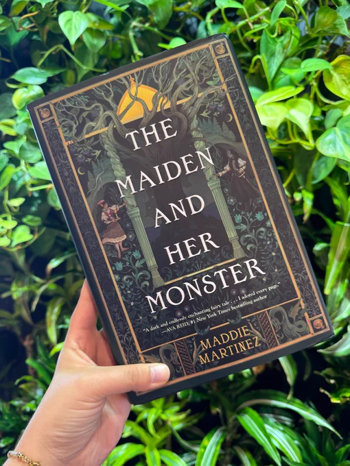 Someone holding up a copy of The Maiden and Her Monster by Maddie Martinez in front of a bunch of green, leafy plants.