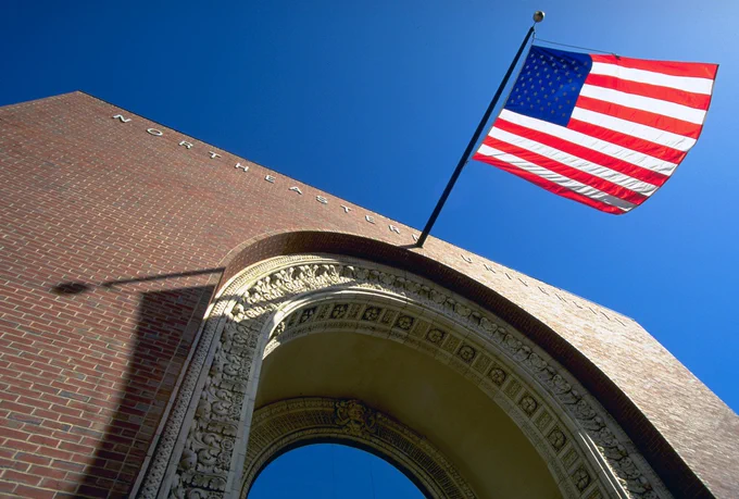 Brick archway in front of a blue sky with an American flag blowing on a pole above it. "Northeastern University" is lettered above it.