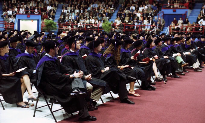 A crowd of students in graduation regalia with purple hoods sit in chairs