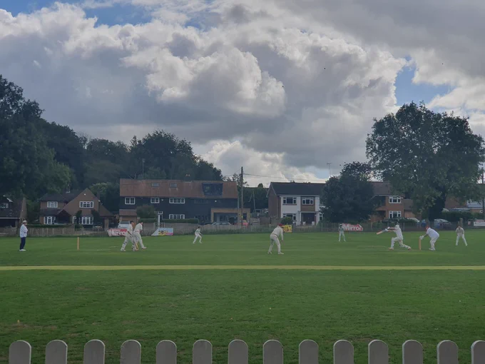 View from the boundary at Farningham shows batter hitting ball as fielders and the umpire looks on. Houses under clouds can be seen in the background and the top of a white fence in foreground