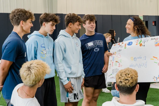 A group of Bethpage High School students stand in a semicircle with an adult mentor during Sources of Strength peer leader training. The students smile as they present a colorful poster filled with drawings and words, while the mentor holds the poster and laughs with them.