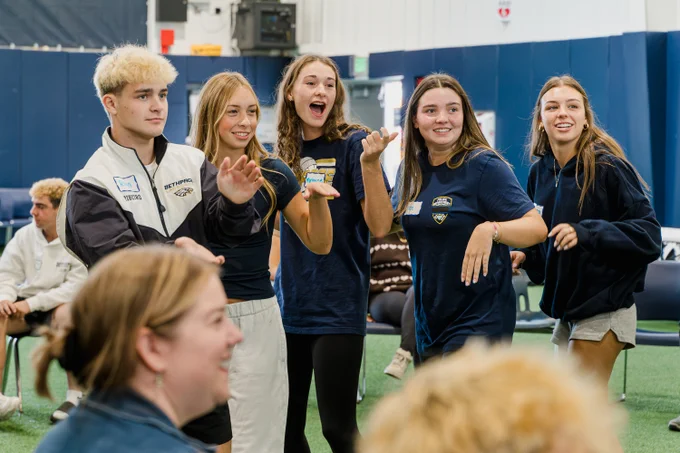A group of female students smile and laugh while participating in the same movement exercise.