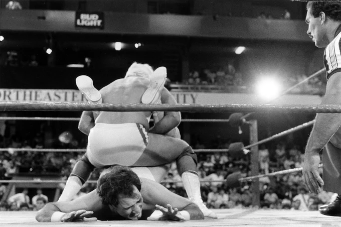 Black and white image of a professional wrestling ring. A man with his back to the camera squats and holds the legs of another man laying on the mat. "Northeastern University" is visible on a wall in the background.