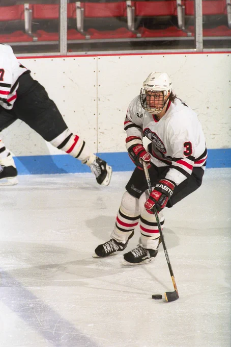 A men's hockey player wearing a white Northeastern University jersey skates on ice in front of a white board.