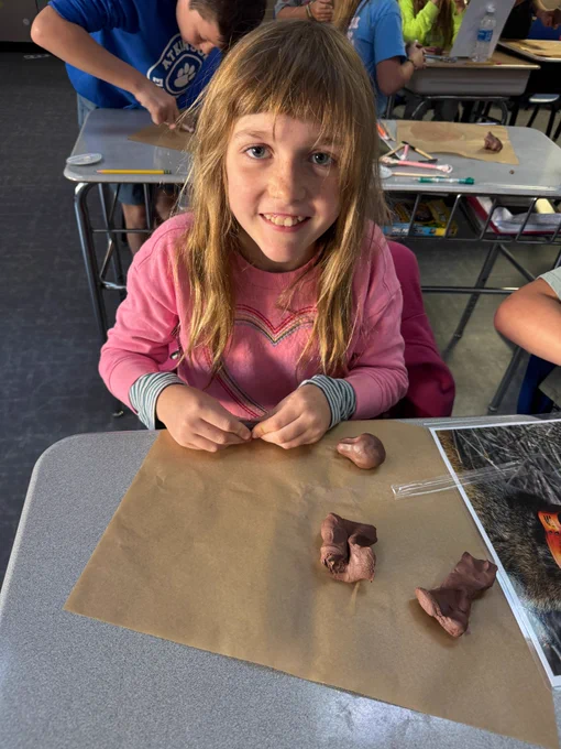 girl with bangs and a pink sweatshirt with a heart on it smiles up at the camera. on her desk in front of her is a red wolf sculpture she made out of red clay.