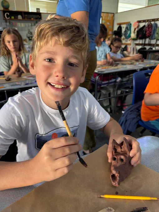 boy in white shirt with blonde hair sits at his desk. kids and a teacher are in the background out of focus. on his desk he has a clay sculpture of a red wolf that he has created and in his hand he has a carving tool