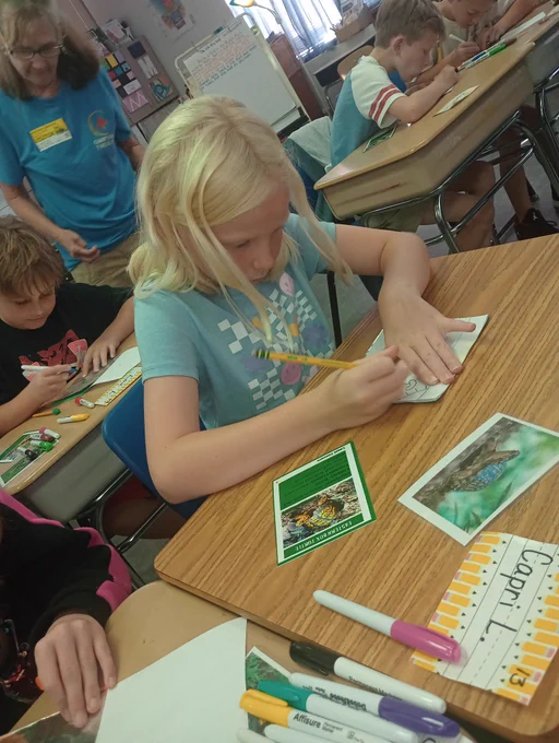girl in a blue shirt with blonde hair hunches over her desk and writes on a small piece of paper with a pencil
