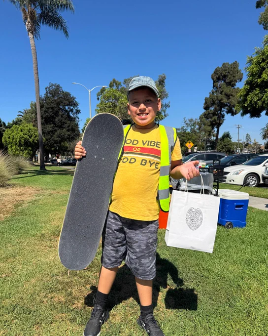A child volunteer holding a skateboard in one hand and a prize bag in the other, standing in a sunny park with cars and trees in the background.