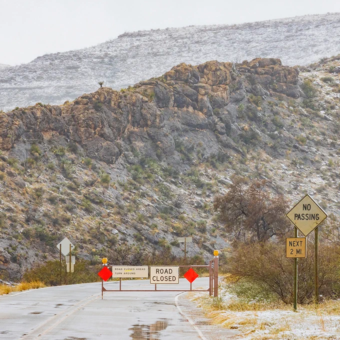 A snowy scene with "Road Closed" and "No Passing" signs on a wet road leading towards a hilly landscape.