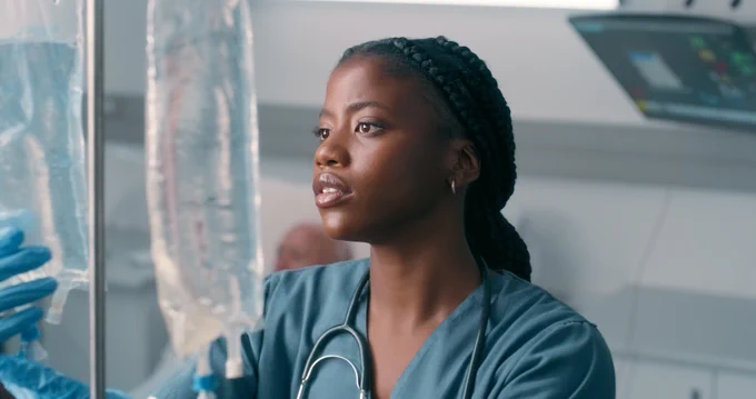 A healthcare worker wearing scrubs and gloves adjusts an IV bag.