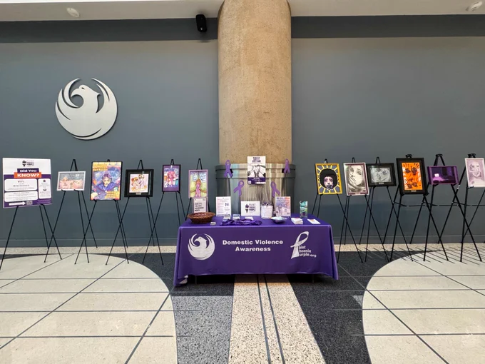 An information booth for Domestic Violence Awareness month, featuring a purple-covered table with promotional materials. Surrounded by posters on easels and a large City of Phoenix logo on the wall behind.