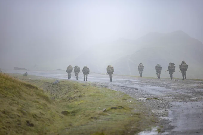 Nine soldiers carrying heavy backpacks walking along a misty, gravel road in a mountainous area.