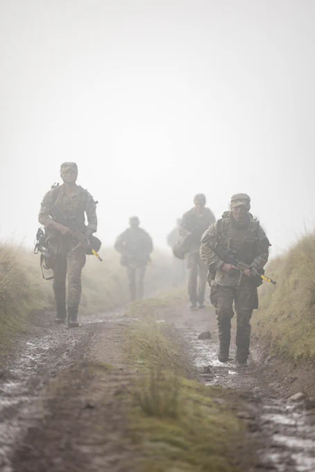 Soldiers in full combat gear walking along a muddy trail through foggy terrain with grass on both sides.