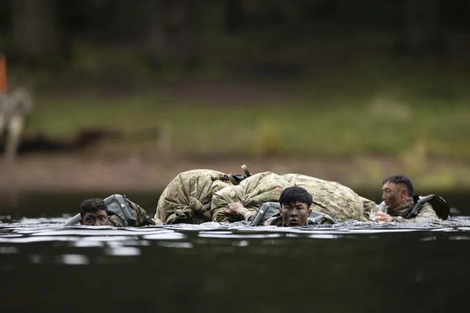 Soldiers in camouflage uniforms wading through water carrying a comrade on their backs during a training exercise.