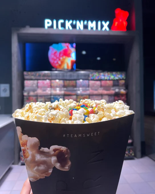 A person holding a popcorn container with "#TEAMSWEET" printed on it, in front of a Pick'n'Mix candy station.