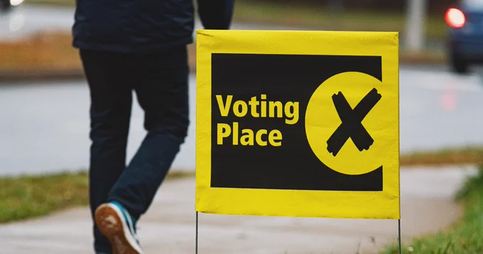A person walking past a yellow "Voting Place" sign with a large black X, located beside a sidewalk.