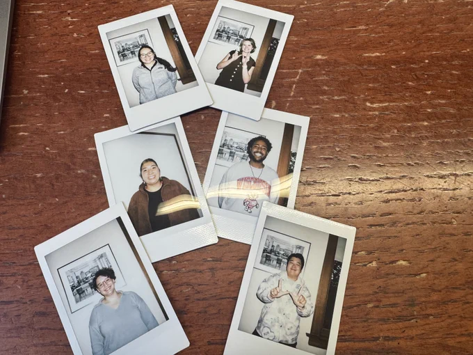 Six Polaroid pictures lay out on a brown, wooden table. Each image features one first-year PhD student. Four students are smiling in their photo, while two are making a Wisconsin "W" with their hands.