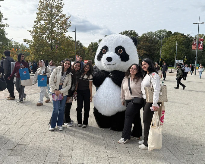 Group of students posing outdoors with a person in a large panda costume on a paved area with trees in the background.