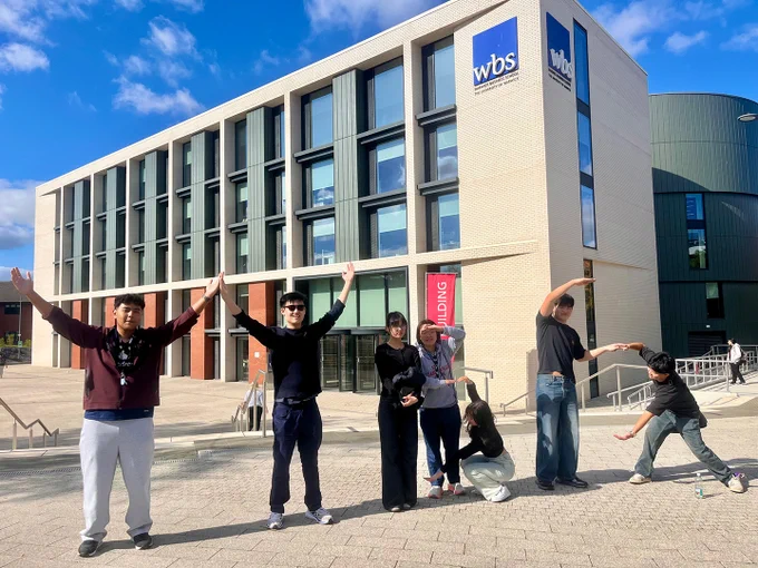 Group of seven people posing in front of the Warwick Business School building on a sunny day, the letters W B S with their arms and bodies.