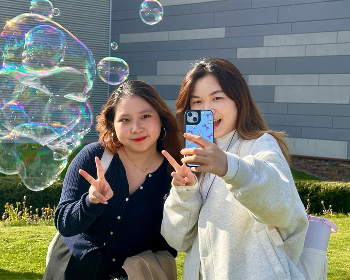 Students outdoors taking a selfie with a smartphone, both showing peace signs with their hands on a sunny day.