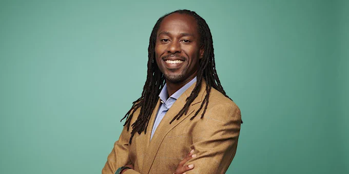 Professor Keon West smiling, wearing a tan blazer over a light blue shirt, standing against a plain teal background.