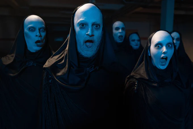 Robert Webb, David Mitchell, Kiell Smith-Bynoe and Krystal Evans in a group of people all with pale faces, dressed in black, hooded robes, and staring into the camera with their mouths wide open.