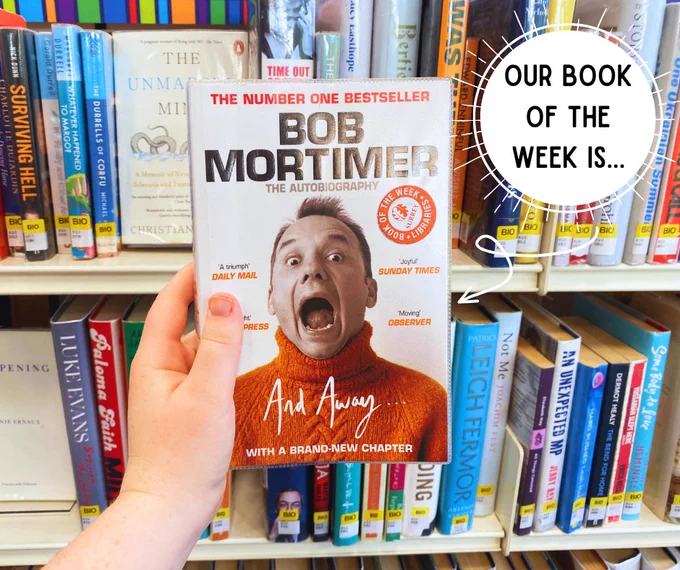 A photo of Bob Mortimer’s book ‘And Away’ in front of a shelf full of colourful books. A white circle with the text, ‘Our book of the week is...’ is in the top right corner. 
