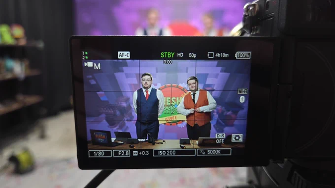A photo of the livestream set at Cubed! 2025.

Taking up most of the frame is the screen of a digital camera, through which Adam and Nath (in their costumes as Jimmy McGee and Dennis Dee) are seen standing behind a desk. They are stood in front of a screen with the Gameshow Showdown logo on it.