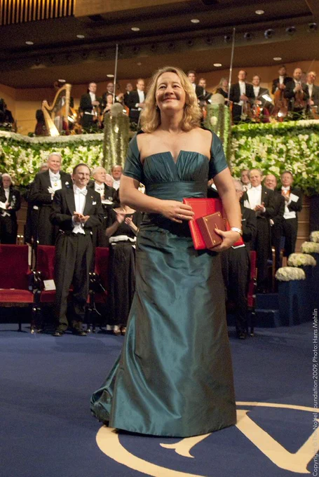 Carol Greider receiving on stage at the Nobel Prize ceremony, dressed in an elegant teal gown, smiling and holding her medal and diploma, rows of applauding laureates stand behind her in formal attire.