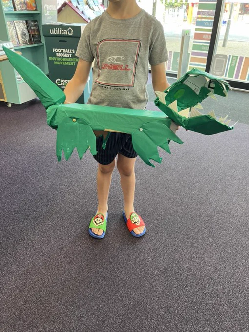 Photo of a little boy wearing black shots and a grey t-shirt holding a green creature made out of cardboard 