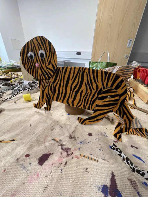 Photo of an animal made out of cardboard and stripey paper - it has googly eyes and a pink nose. You can see craft materials in the background