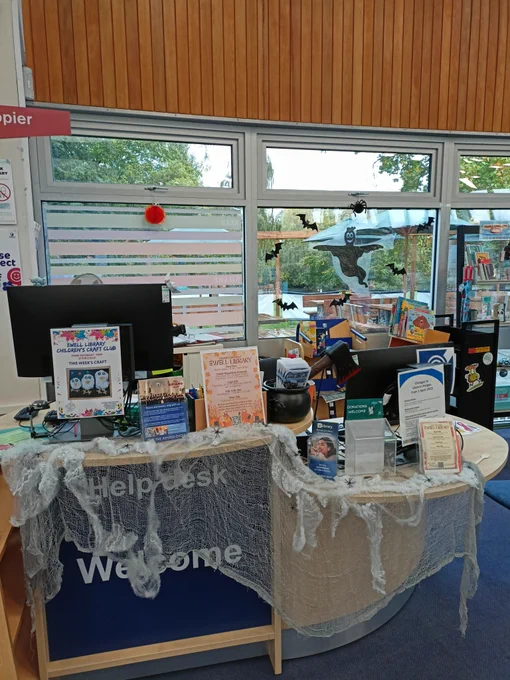 Photograph of a wooden desk with two computers, information posters, and a small plastic cauldron on it. The front of the desk is decorated with grey gauze resembling cobwebs with small plastic spiders stuck on it.