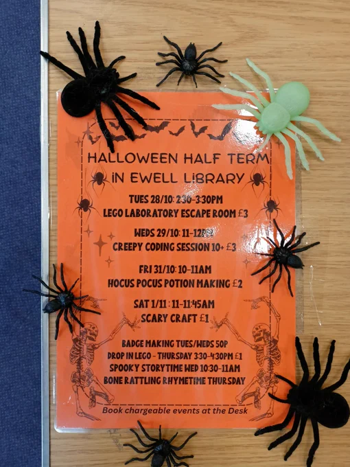 Photograph of an orange activities flyer taped to a wooden table with plastic spiders scattered around it. Events include a Lego Laboratory Escape Room on Tuesday 28th at 2:30, a Creepy Coding Session on Wednesday 29th at 11:00, and Hocus Pocus Potion Making on Friday 31st at 10:00. 