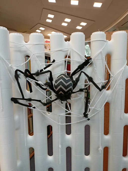 Photograph of a large fake spider hanging from a white wool web strung across a white foam wall. 