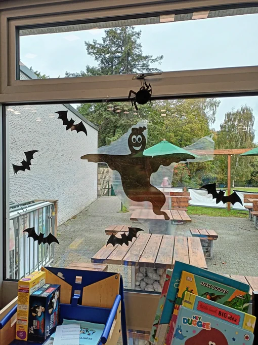 Photograph of a window with a black plastic ghost silhouette stuck to it. The ghost is surrounded by black paper bats, and there is a bat paper lantern hanging from the window handle.