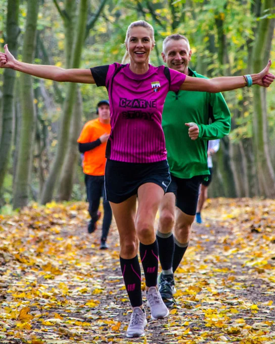 A parkunner running through autumnal brown leaves with her arms out doing thumbs up.