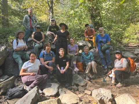 14 people gather for a group photo in an area with boulders near the stream at Rock Creek park