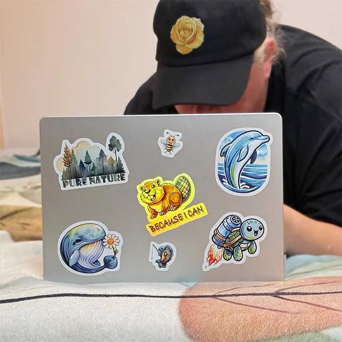 A silver laptop is decorated with colorful stickers, including a dolphin, a beaver with the text Because I can, a whale holding a flower, a forest with Pure Nature, a rocket turtle, a bee and a small fish. A person wearing a black cap with a yellow rose emblem is leaning over the laptop. The stickers are bright, playful and creative, ideal for customizing personal items.