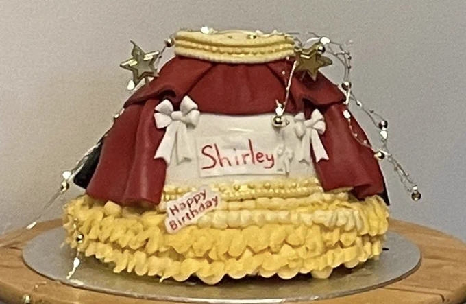 A cake decorated with red curtains and decorated to look like a stage. It says Happy Birthday Shirley. Highlight decorations in gold with white bows